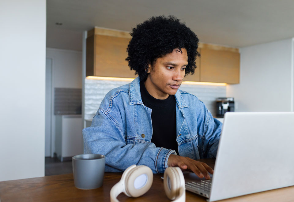 Person wearing a jean jacket works at a laptop with headphones resting on the table.