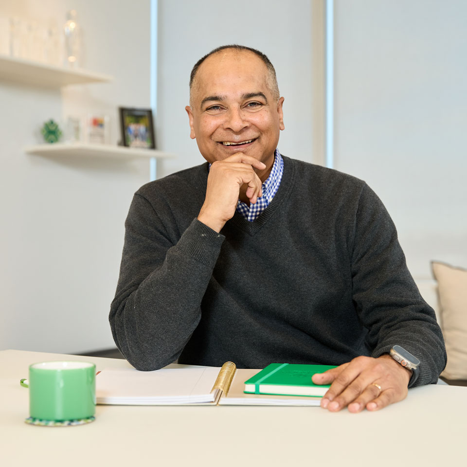 Middle aged person smiles while seated at a desk with a green covered notebook.