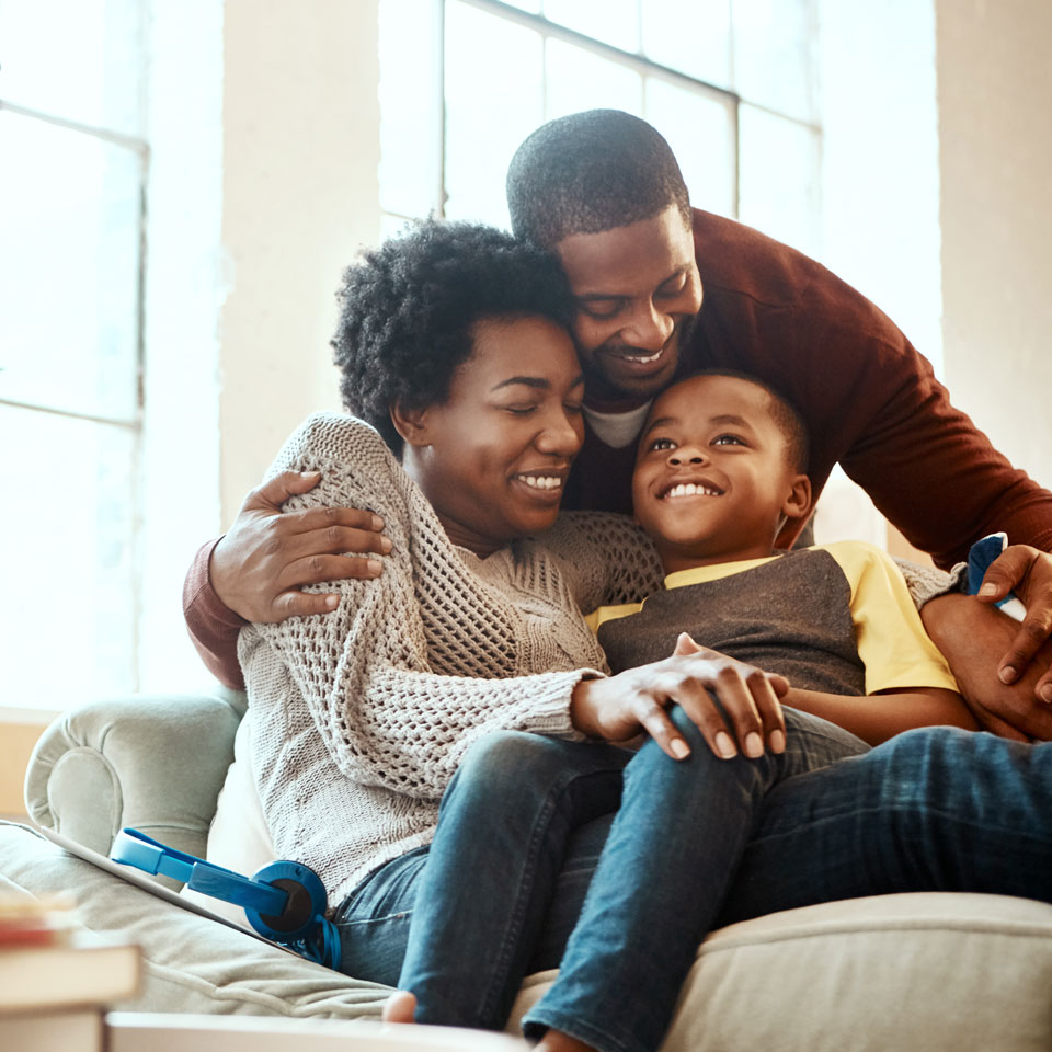 Two adults and young child embrace one another on a couch, smiling.