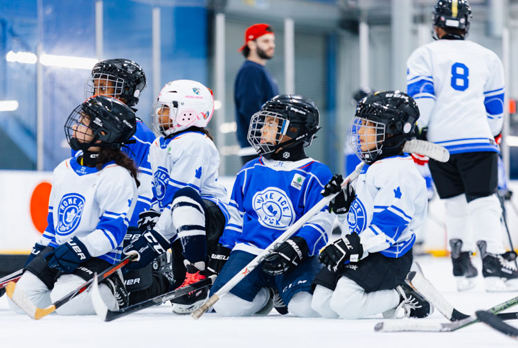 Group of children in hockey equipment sit on the ice at a hockey rink.