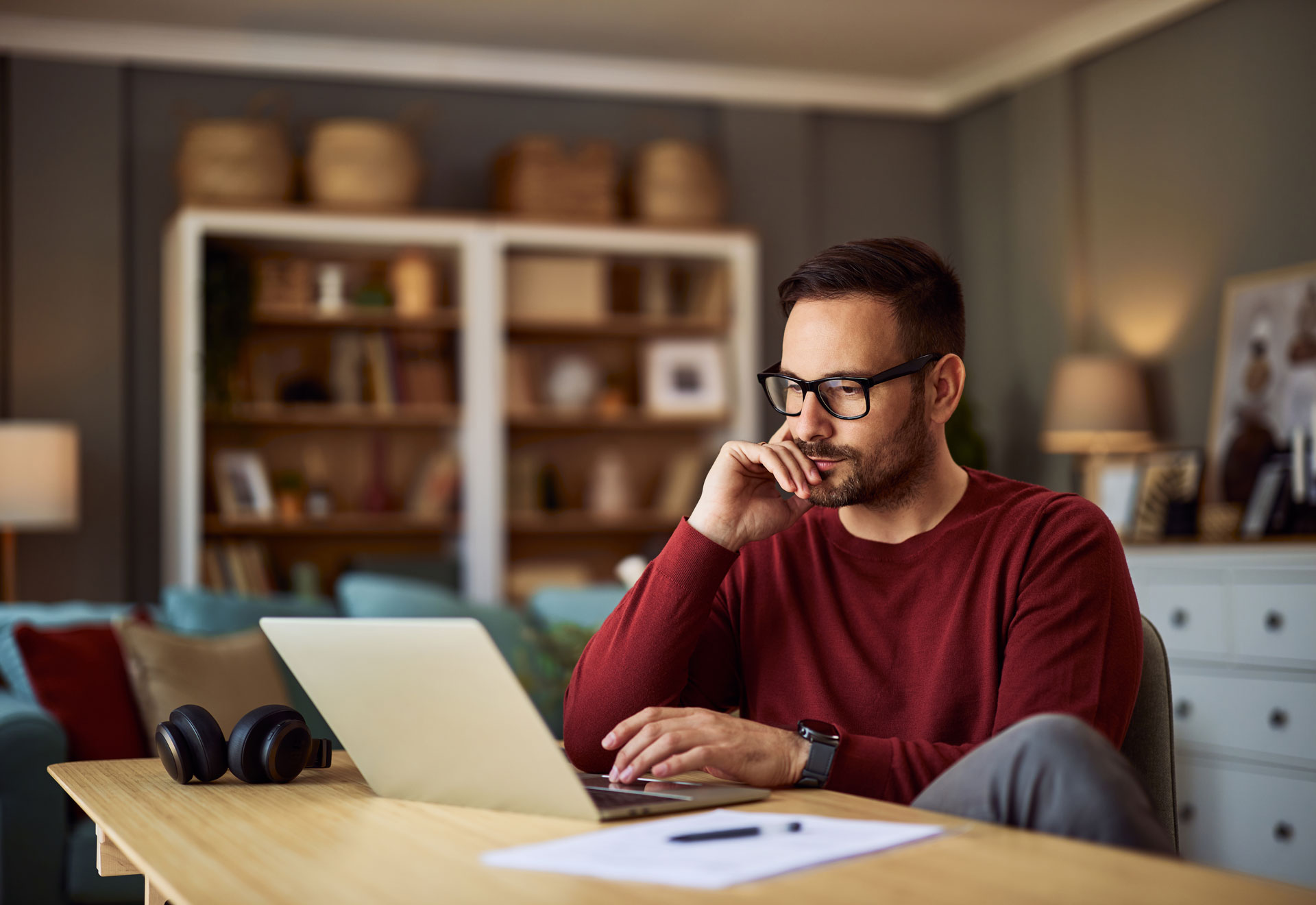 A person with glasses and a beard works on a laptop in a home office.