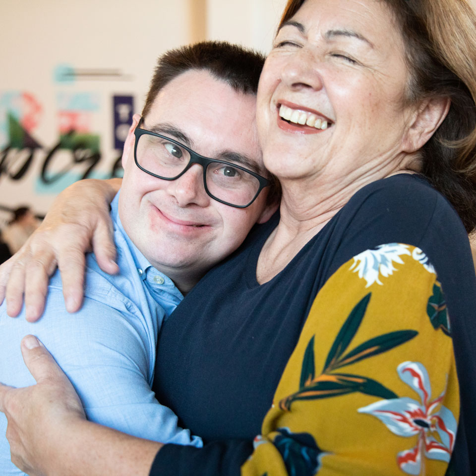 Young adult in glasses and an older person smile as they hug one another.