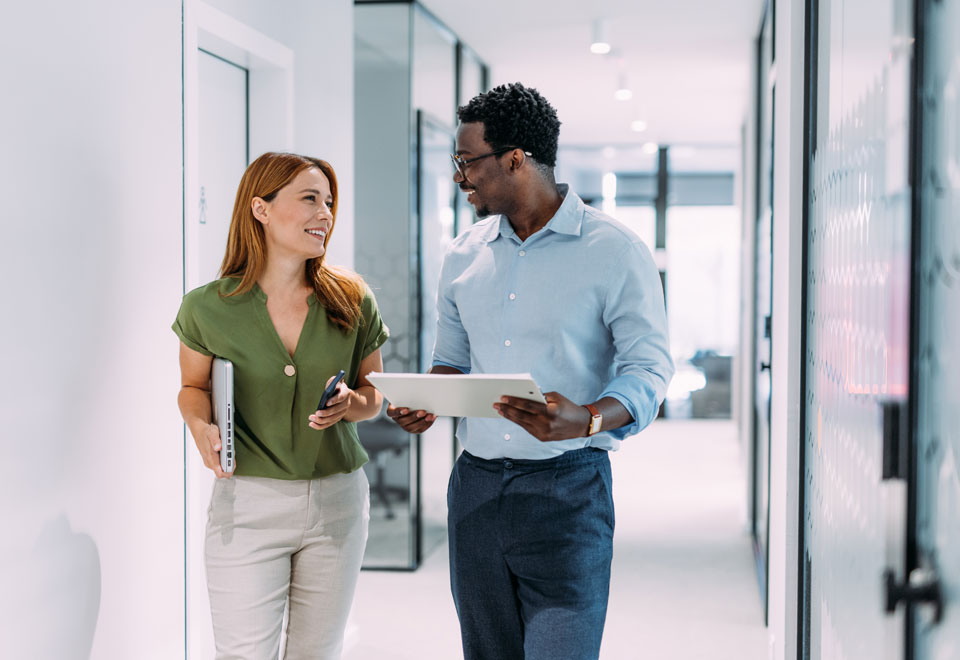 Two professional people chat while walking in an office hallway.