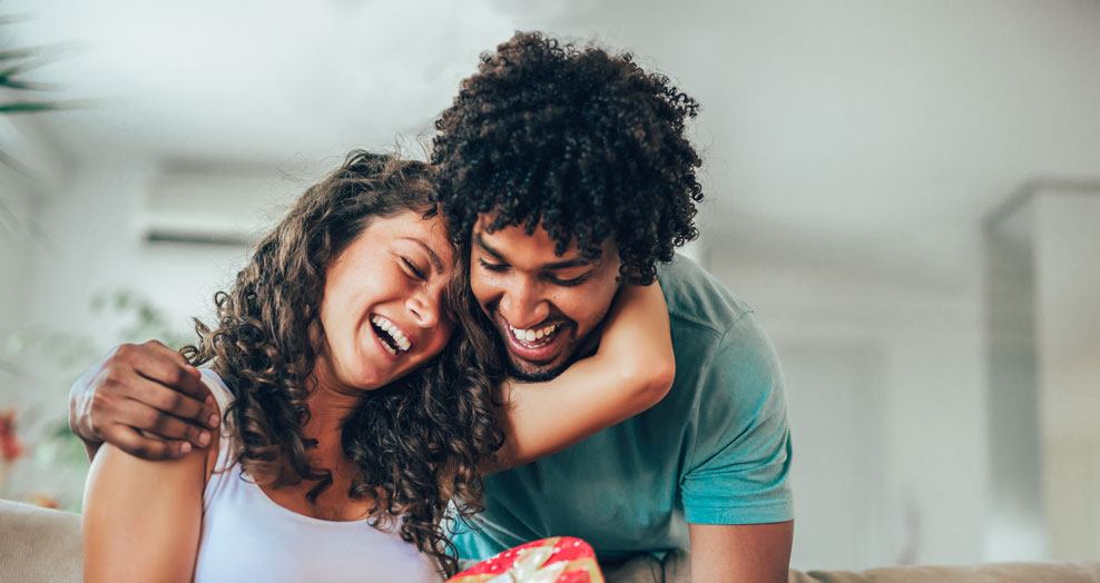 A young adult couple smile and embrace while exchanging a gift.