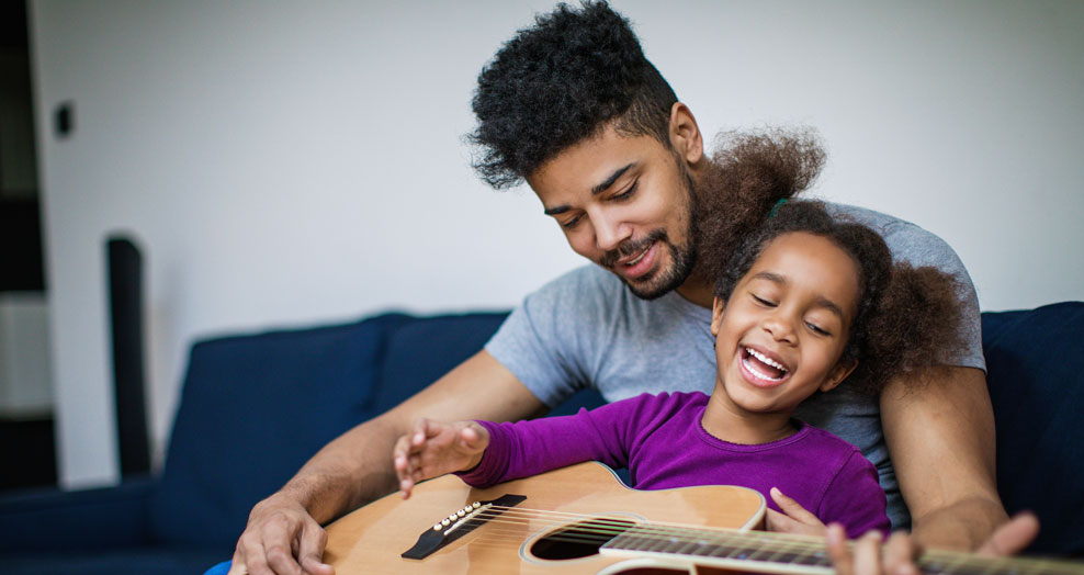 Young child and a parent sit together smiling while holding a guitar.