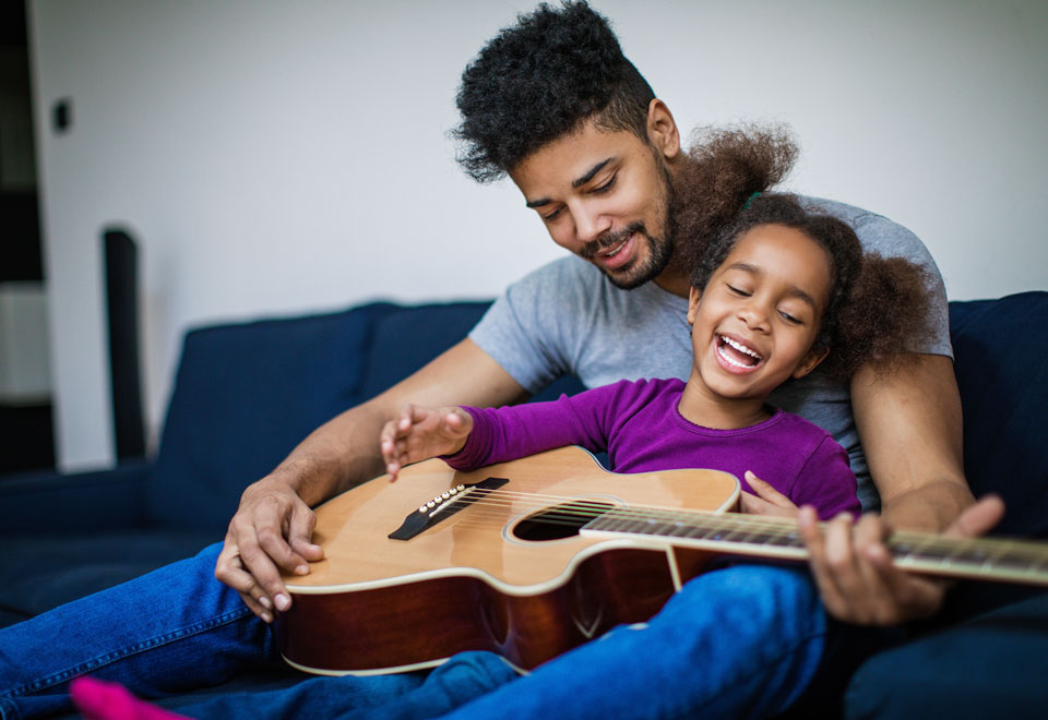 Young child and an adult sit together smiling while holding a guitar.