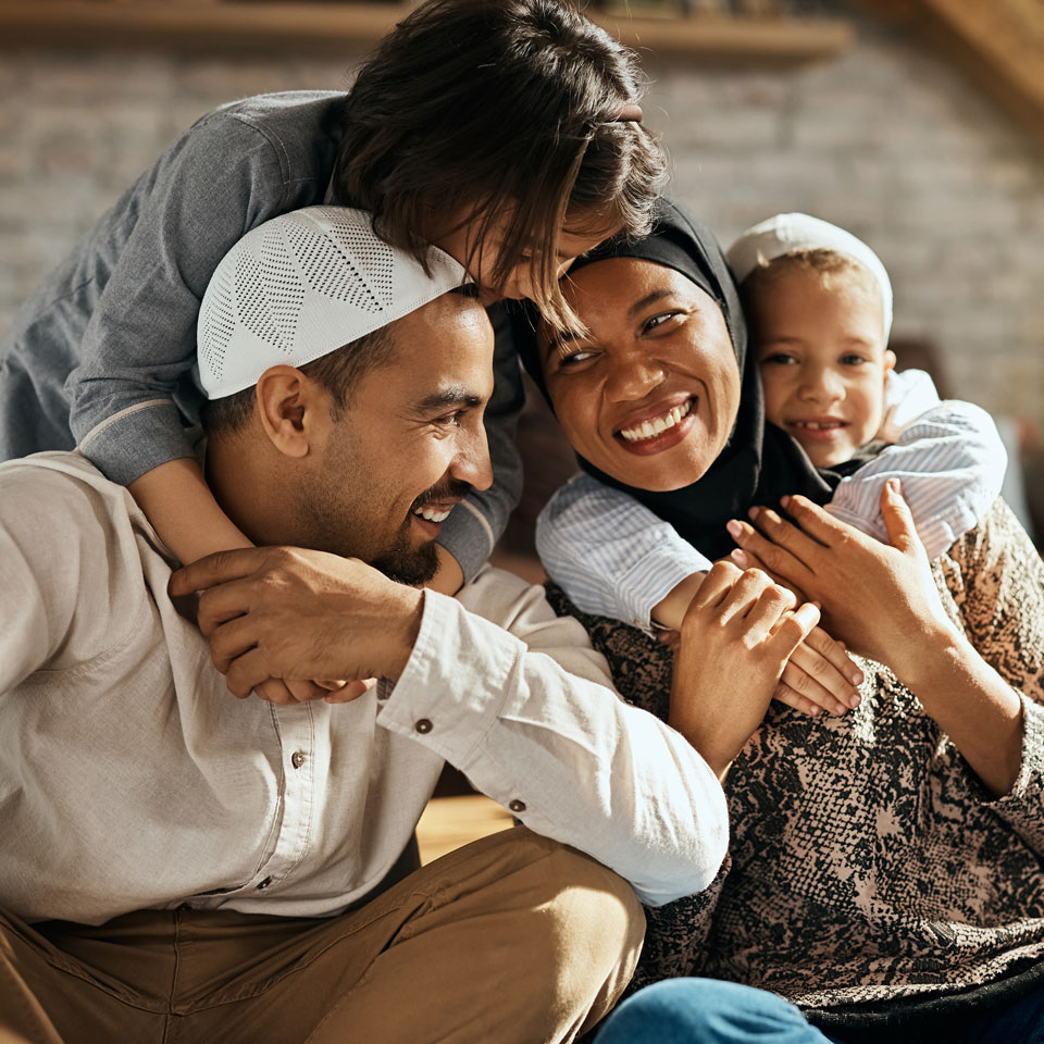 Young family wearing hijabs and taqiyahs sit and smile together.