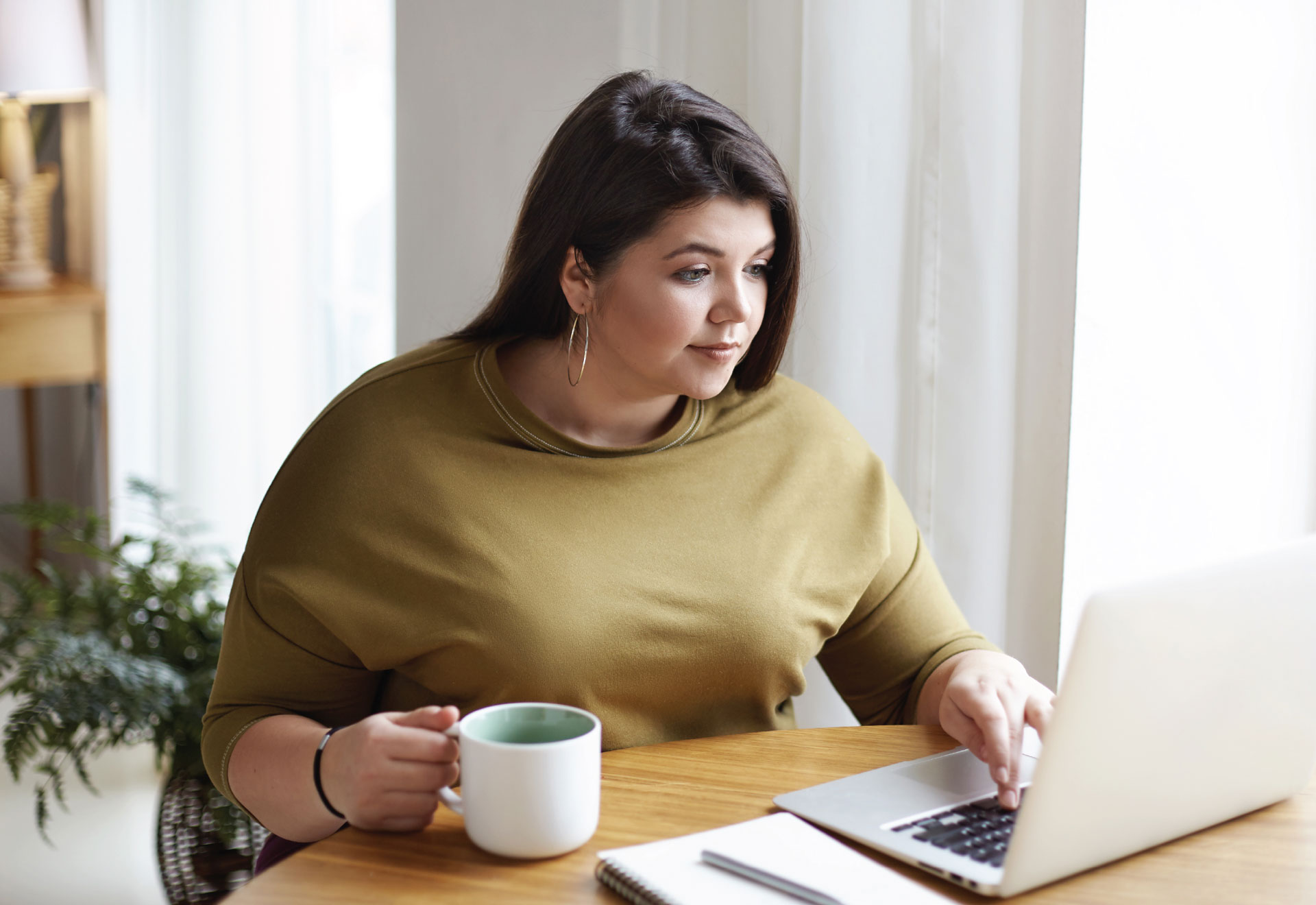 Person with long hair sits at a table with a coffee and an open laptop.