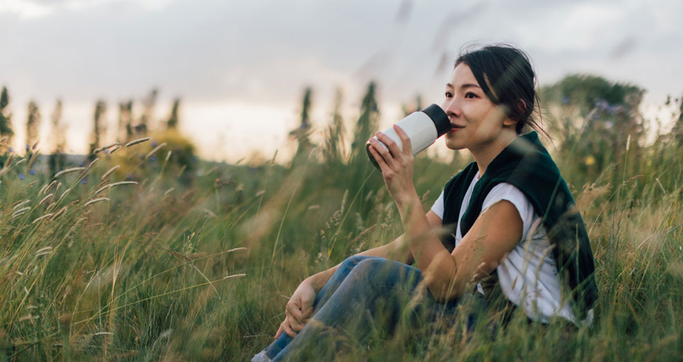 Person with long hair sitting in a field drinking from a water bottle.