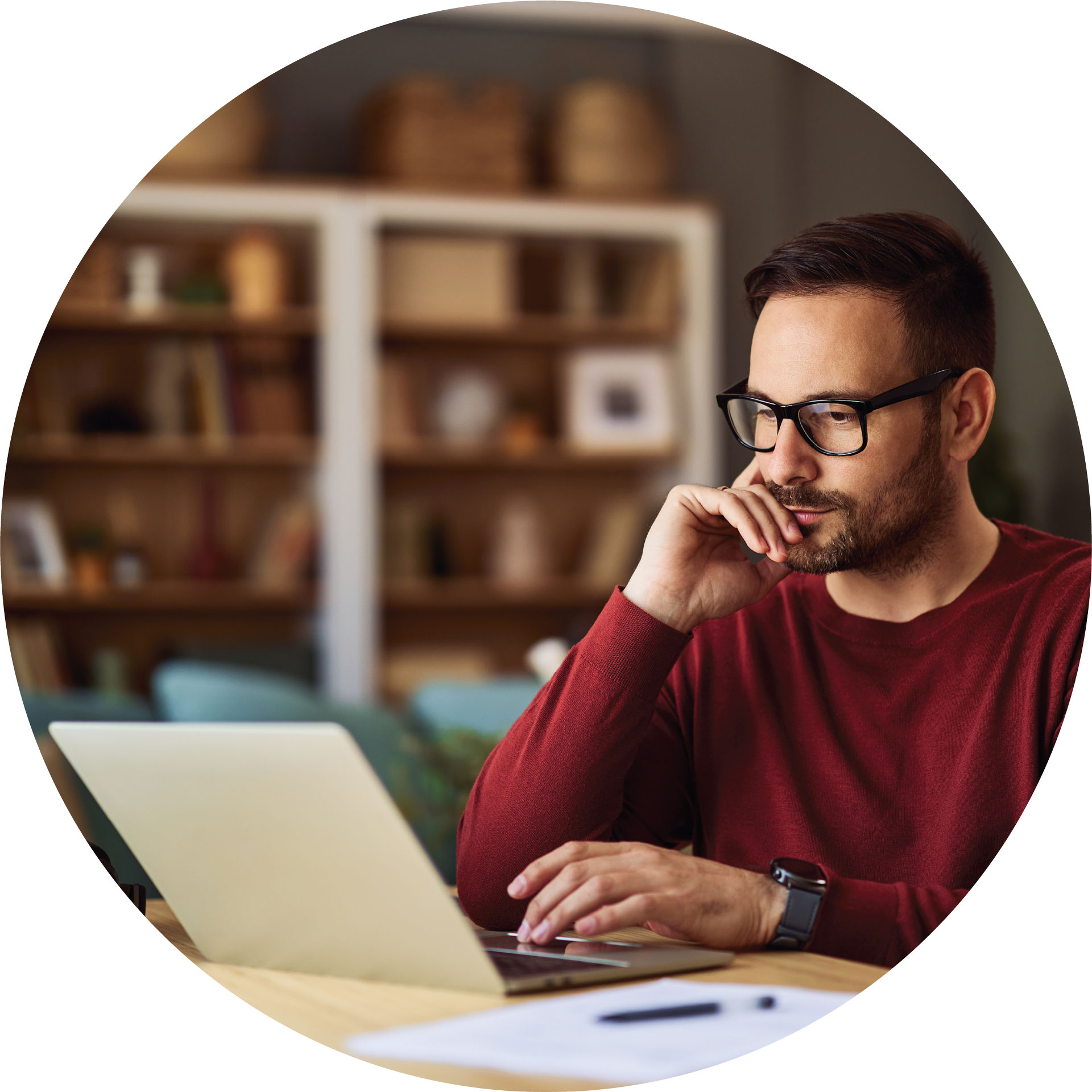 Person with glasses and a beard works on a laptop in a home office.