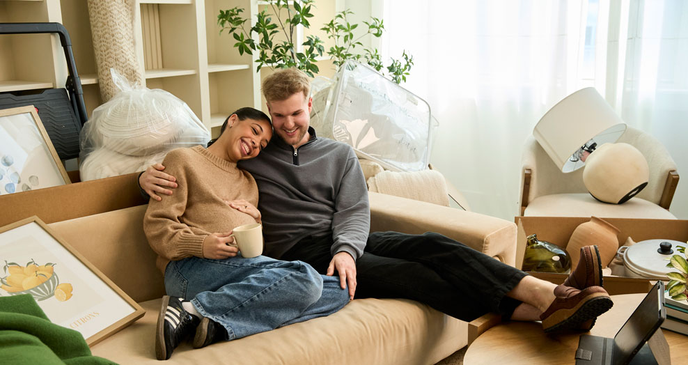 Pregnant person and their partner sit on a couch surrounded by moving boxes.