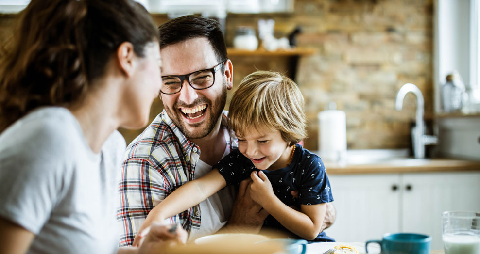 Parents and a young child laugh together in a kitchen.