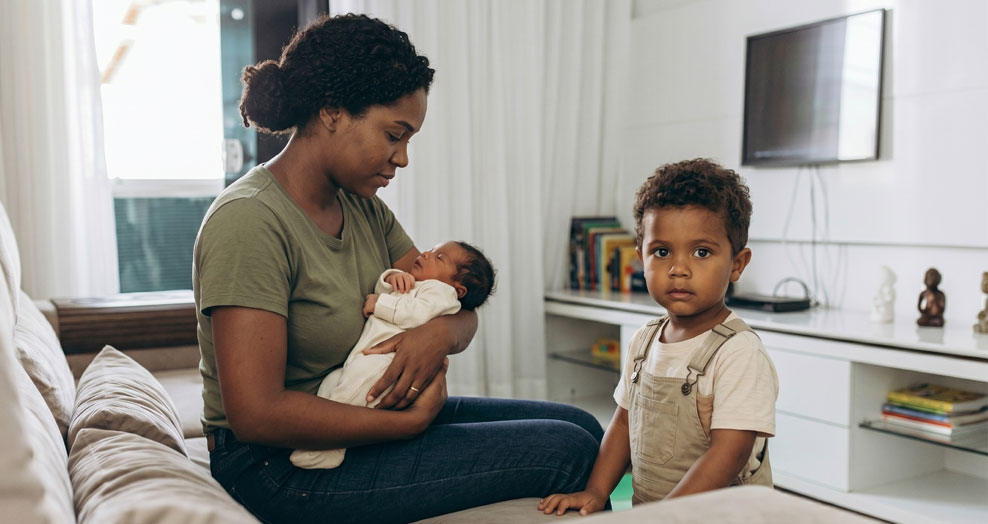An adult cradles a baby in a living room while a toddler looks on.