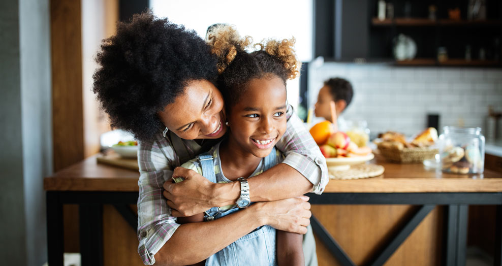 Parent hugs a smiling child in a kitchen.