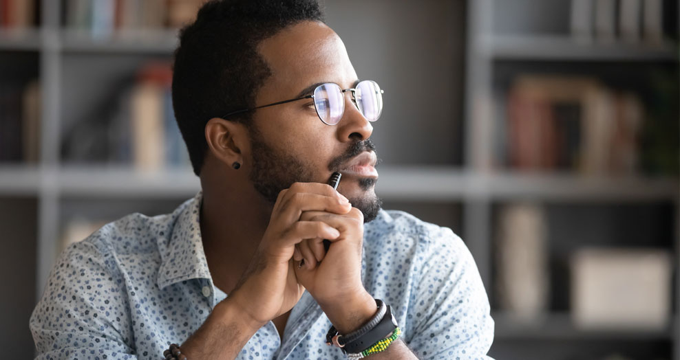 Person with glasses holds a pen while looking toward a window.