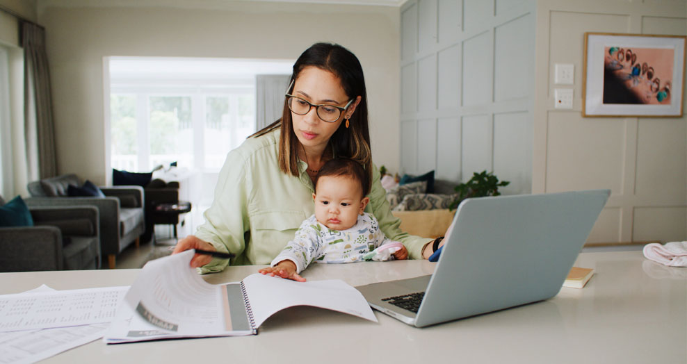 Person works at a desk with a laptop and a notebook with a baby on their lap.