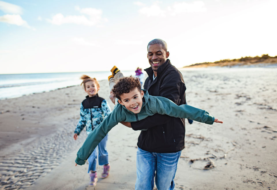 Parent carries a young child on a beach who pretends they're flying, while another child looks on.