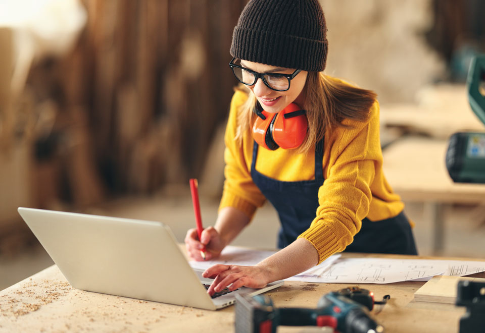 Contractor with earmuffs around her neck stands at a table with an open laptop and a drill.