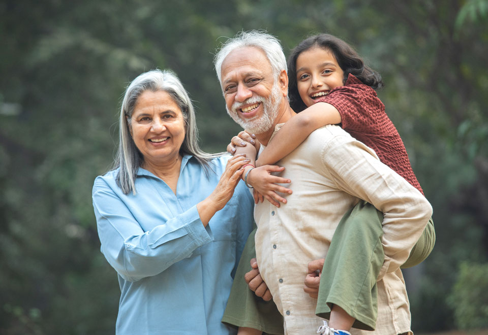 An elderly person standing beside their partner gives a piggyback ride to a child.