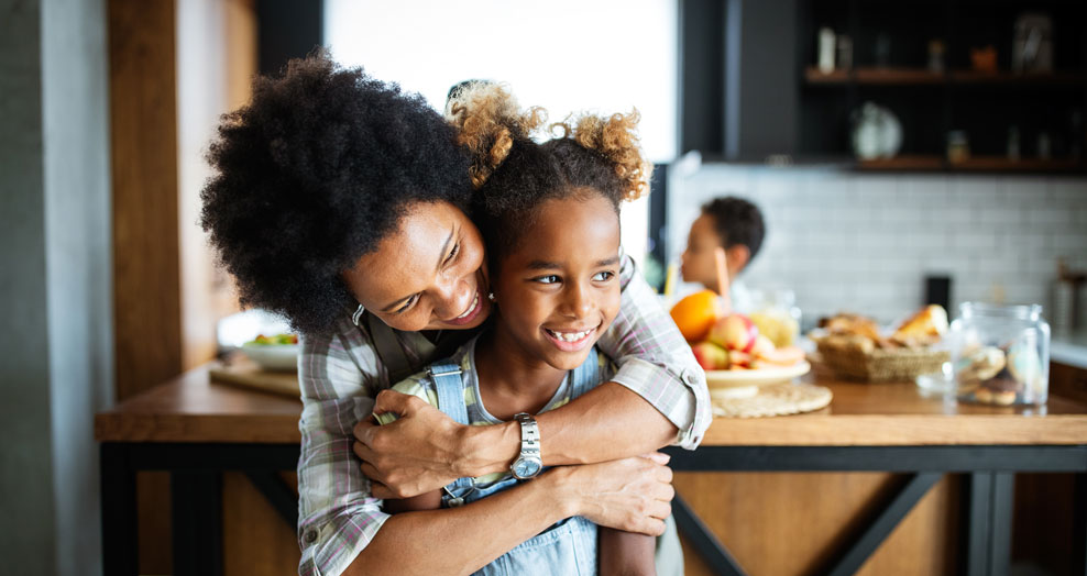 Parent hugs a smiling child in a kitchen.