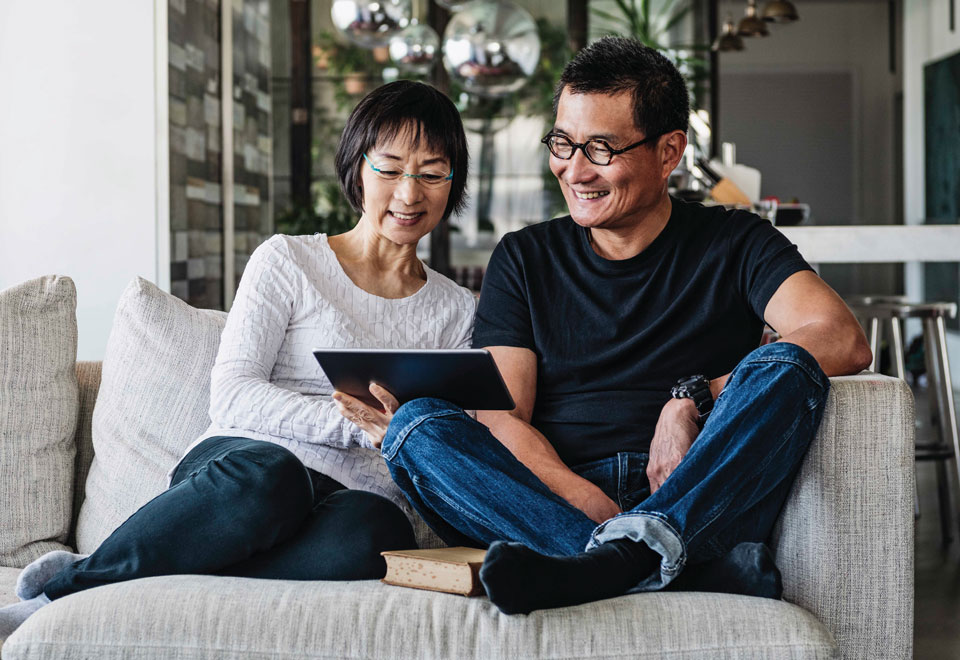 Couple sits together on a couch smiling while looking at a tablet.